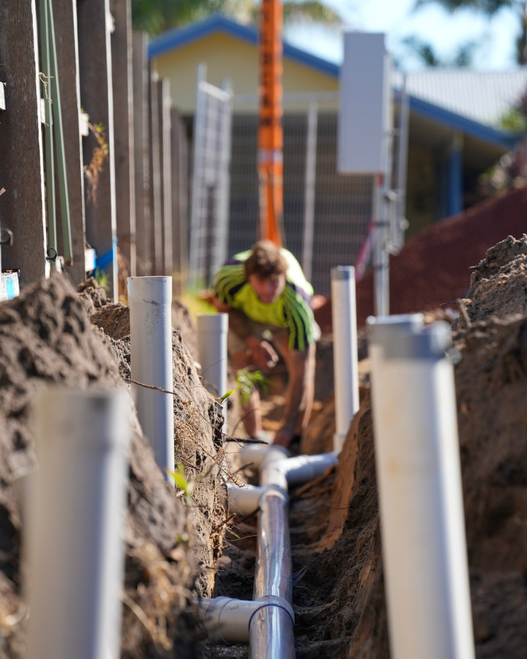 Long backyard trench with a row of vertical PVC stormwater stacks set in fresh sand, apprentice working in the background.