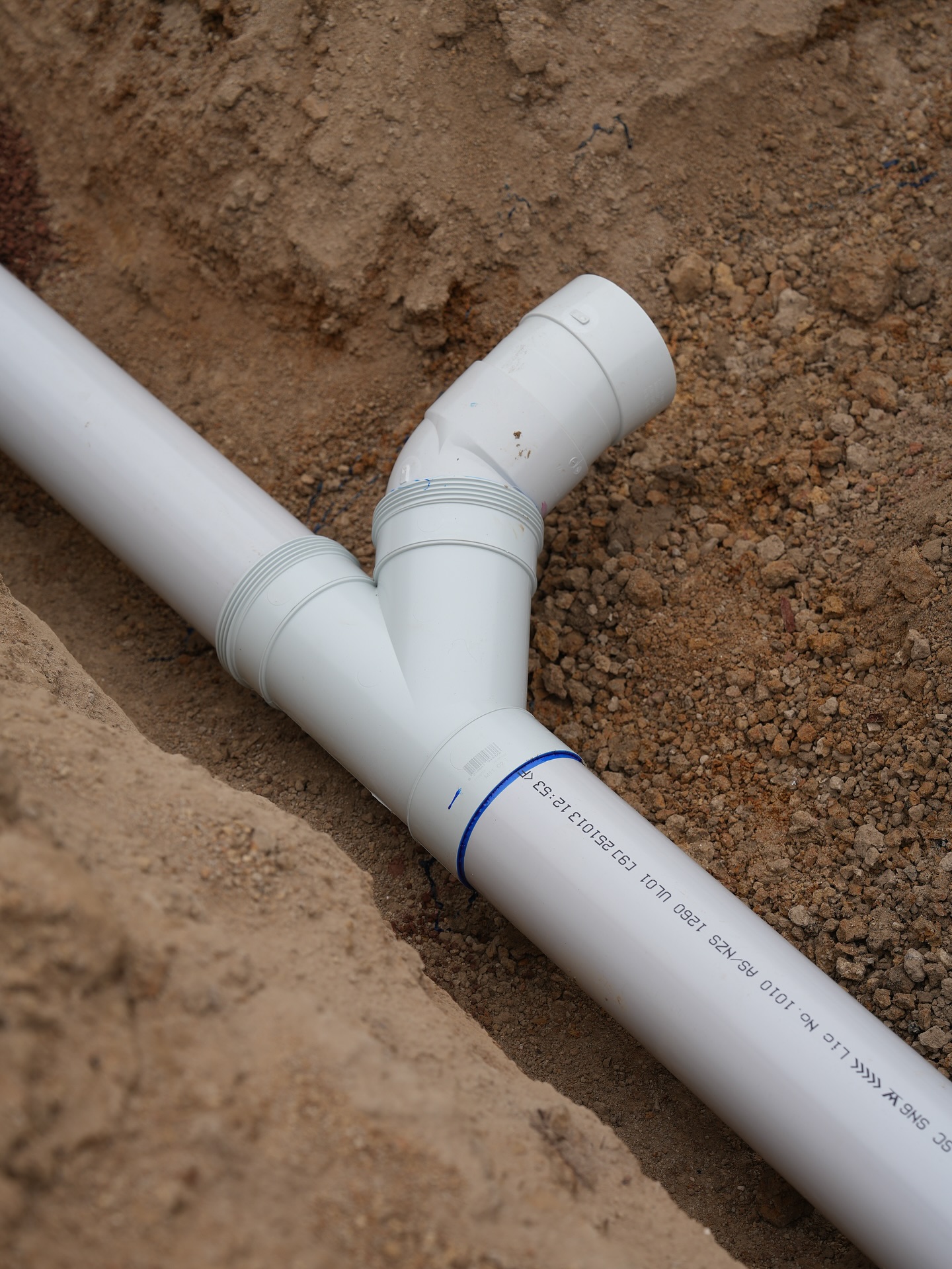 Close-up of a DWV sewer junction seated in a trench, fresh white PVC against orange dirt.