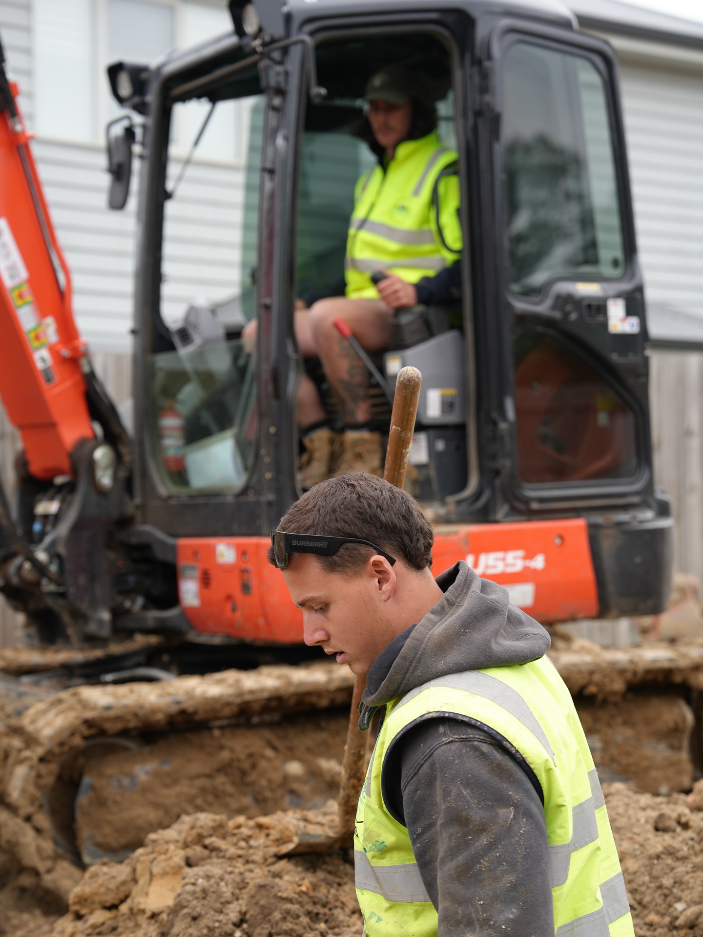 Orange Kubota U55-4 excavator with operator in the cab. A second crew member in branded hi-vis works in the foreground.