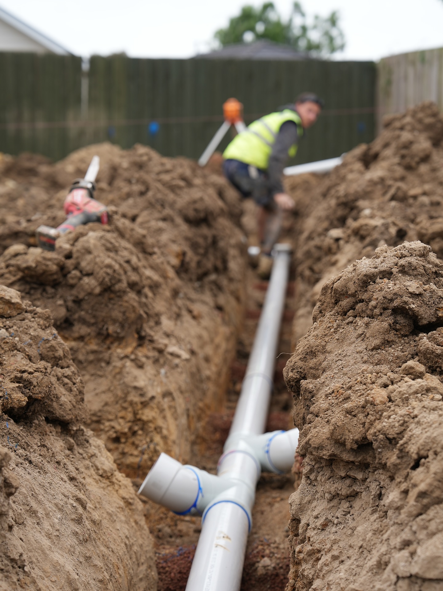 Deep narrow trench with a fresh PVC sewer junction at the base. A leading hand in hi-vis stands above measuring fall.